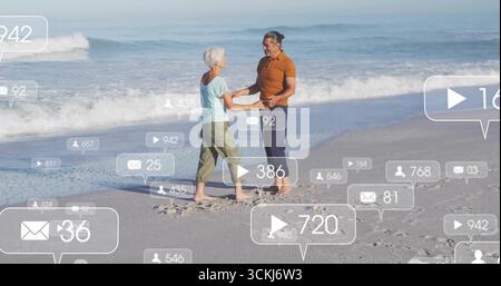 Lächelndes Seniorenpaar, das am Sandstrand am Wasser steht, mit schwimmenden Social-Media-Ikonen Stockfoto