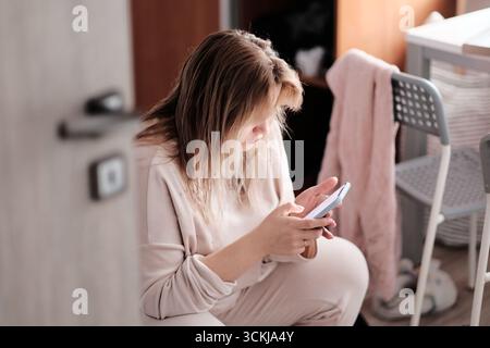 Eine junge Frau sitzt drinnen in lässiger Loungewear, konzentriert sich auf ihr Smartphone, verbringt Zeit online und entspannt sich zu Hause. Stockfoto