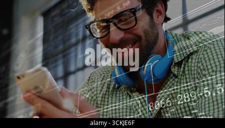 Lächelnder Mann mit Brille und blauen Kopfhörern, der Smartphone am Arbeitsplatz analysiert, Daten-Overlays Stockfoto