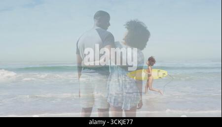 Ein Paar, das am Sandstrand steht, mit einem laufenden Surfer im Badeanzug und gelbem Surfbrett Stockfoto