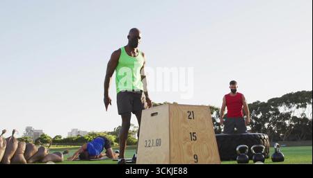 Mann in grünem Tank neben Plyometriekasten auf Parkrasen, mit Fitnessgeräten Stockfoto