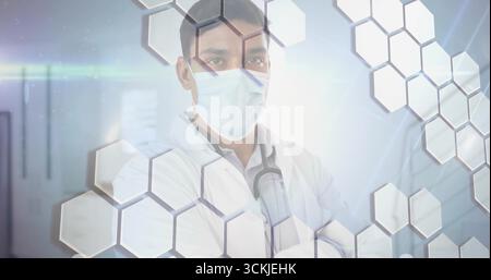 Arzt stehend mit Laborkittel, chirurgischer Maske im Kliniksaal, mit Stethoskop, Sechskant-Overlays Stockfoto