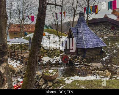 Ein charmantes Holzhaus mit blauem Dach steht neben einem kleinen Bach. In der Nähe hängen bunte Fahnen, die einen Kontrast zum schneebedeckten Boden und dem Winter bilden Stockfoto