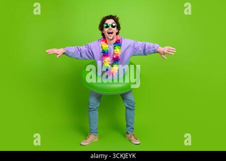 Energiegeladener junger Mann in farbenfrohem Outfit mit tropischem Flair auf lebendigem grünem Hintergrund, der die Arme in verspielter Pose weit hält Stockfoto
