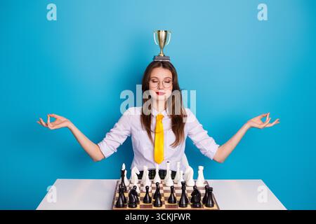 Kluge Frau meditiert mit Trophäe auf dem Kopf, die am Schachbrett sitzt und Fokus und Leistung gegen Blau symbolisiert Stockfoto