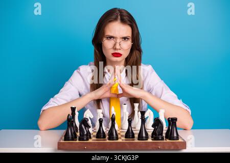 Junge Frau in Brille konzentriert sich auf Schachspielstrategie mit Selbstvertrauen vor einem leuchtend blauen Hintergrund Stockfoto
