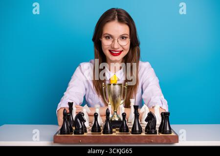 Selbstbewusste junge Frau feiert den Schachsieg mit einer Trophäe vor hellblauem Hintergrund Stockfoto