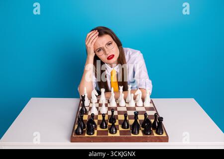 Junge brünette Frau im Retro-Outfit denkt während intensivem Schachspiel mit leuchtendem blauem Hintergrund Stockfoto