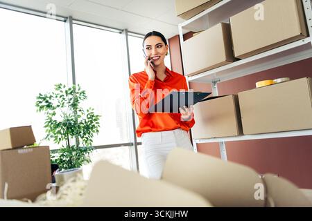 Junger Unternehmer, der den Produktbestand verwaltet, während er telefonische Bestellungen in einem modernen Büro mit großen Fenstern entgegennimmt Stockfoto