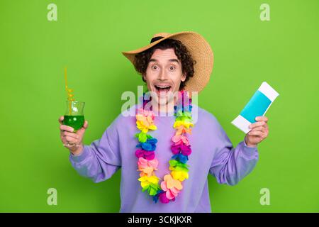 Fröhlicher junger Mann mit farbenfrohen lei und Strohhut, der Getränke und Tickets hält und Begeisterung für Reisen zeigt Stockfoto