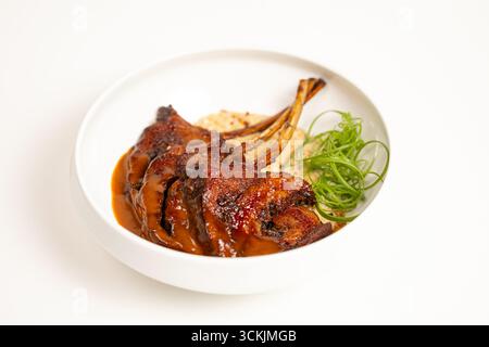 Saftige gegrillte Lammkoteletts mit kräftiger brauner Sauce, serviert mit cremiger Beilage und Kräutern, fotografiert auf weißem Hintergrund für Food Design Projekte. Stockfoto