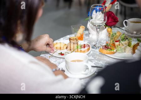Eine Frau genießt elegante Häppchen auf einem festlichen Tisch mit Tee, Wein und Blumendekor. Elegante Atmosphäre eines formellen Dinner- oder Hochzeitsempfangs Stockfoto