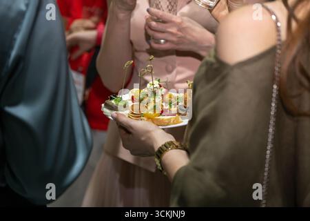 Eine Frau, die einen Teller mit Gourmet-Häppchen bei einer gesellschaftlichen Veranstaltung hält. Gäste genießen Drinks und Vorspeisen in festlicher Atmosphäre. Stockfoto
