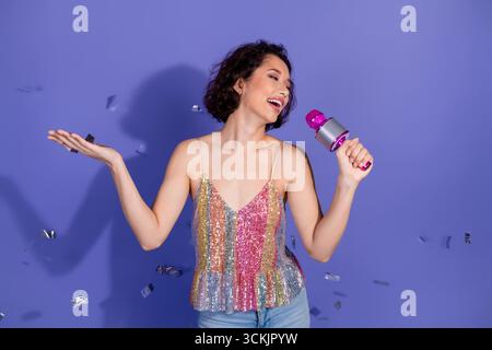 Junge Frau, die mit Mikrofon singt, genießt eine festliche Feier in farbenfroher Kleidung vor einem leuchtend violetten Hintergrund Stockfoto