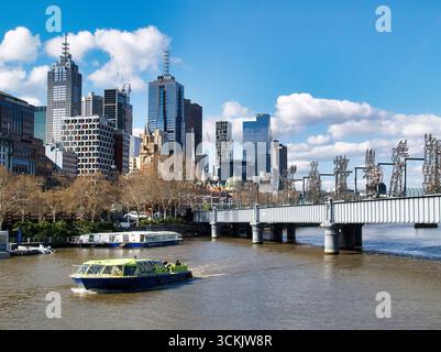 Die Skyline von Melbourne, der Yarra River mit dem Wasserfahrzeug und die Sandridge Fußgänger- und Radbrücke, die einst eine Eisenbahnbrücke war. Stockfoto
