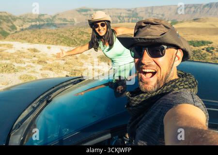 Glückliches junges weißes Paar, das Selfie macht, während es in einem schwarzen Mietwagen sitzt und eine Roadtrip durch Europa macht. Outdoor-Lifestyle-Aktivität auf Roadtrip Stockfoto