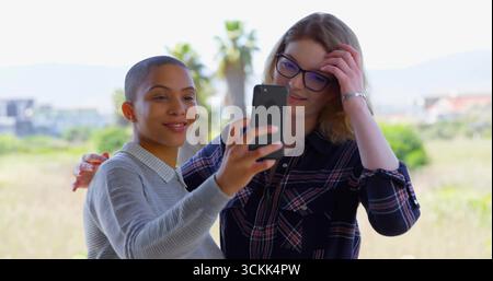 Lächelnde Freunde tragen Pullover und kariertes Hemd, um Selfie auf dem Smartphone auf der sonnigen Terrasse aufzunehmen Stockfoto