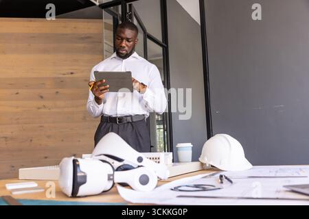 Afroamerikaner, der im Büro steht und Pläne auf einem Tablet mit Bleistift, Helm und VR-Ausrüstung überprüft Stockfoto