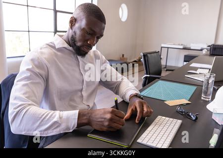 afroamerikanischer Mann in weißem Hemd, der auf Tablette in der Nähe eines Schutzhelms am Schreibtisch skizziert Stockfoto