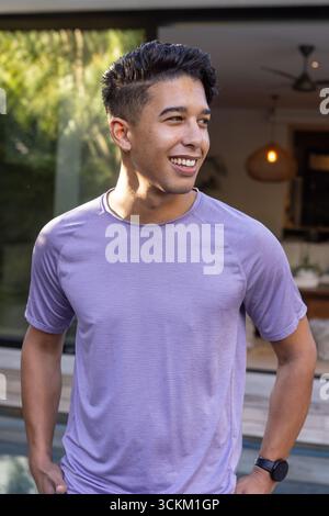 Asiatischer Mann, der auf der Terrasse am Pooldeck steht, Glastüren mit violettem Sportshirt Stockfoto
