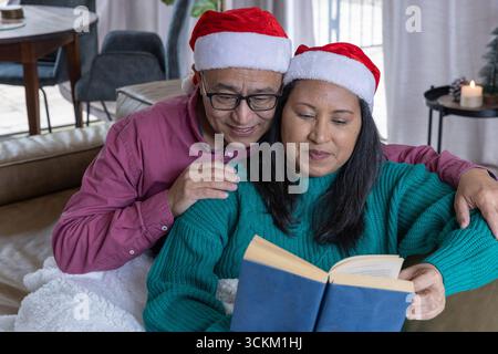 Verschiedene Paare sitzen auf dem Sofa mit weihnachtsmützen und lesen Hardcover-Buch im Wohnzimmer Stockfoto