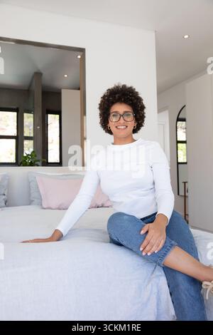 Afroamerikanerin sitzt auf der Bettkante in einem modernen Schlafzimmer mit pastellfarbenen Kissen und Topfpflanzen Stockfoto