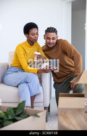 Verschiedene Paare sitzen zu Hause auf dem Sofa und halten eine Kaffeetasse aus Papier, während sie das Smartphone in der Nähe von Boxen betrachten Stockfoto