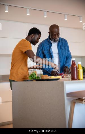 afroamerikanischer Vater und Sohn bereiten Burger auf der Kücheninsel mit Messer, Brötchen, Gewürzen zu Stockfoto