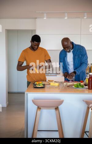 afroamerikanischer Vater und Sohn bereiten Gemüse vor, schneiden Käse auf der Kücheninsel-Theke Stockfoto