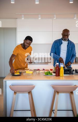 afroamerikaner Vater, Sohn, der auf der Kücheninsel kocht, Gemüse auf Schneidebrett hackt Stockfoto