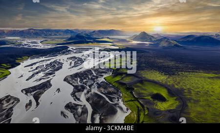 Aus der Vogelperspektive auf Islands geflochtene Flüsse, die durch dunklen vulkanischen Boden fließen, mit grünem Moos und entfernten konischen Bergen unter einem goldenen Sonnenuntergang Stockfoto