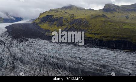 Ein Gletscher in Island mit tiefen Spalten und dunklem Sediment, umgeben von steilen moosbedeckten Klippen unter niedrigen hängenden Wolken in einer stimmungsvollen Atmosphäre. Stockfoto