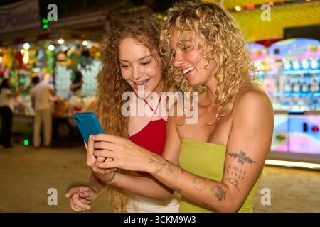 Zwei lächelnde junge Frauen, die nachts im Vergnügungspark ihr Smartphone benutzen Stockfoto