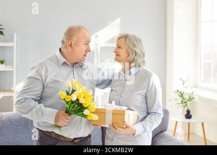 Älterer Mann schenkt Blumen und Überraschungsgeschenk an alte, lächelnde Frau, die eine Geschenkbox hält Stockfoto