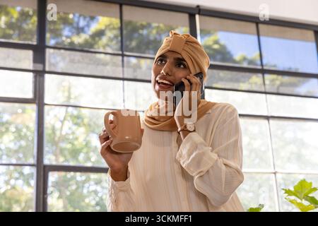 Mittelerwachsene indische Frau, die am Telefon spricht und in einem hellen Büro mit Topfpflanze einen Becher hält Stockfoto