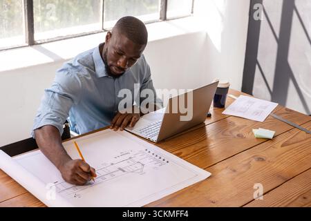 afroamerikaner skizziert mit Bleistift auf Blaupausen am Schreibtisch des Designstudios, Laptop Stockfoto