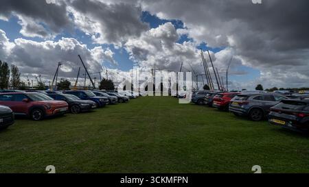 Moderne Autos parkten auf einem grasbewachsenen Feld mit Baukränen, die im Hintergrund unter dramatischem bewölktem Himmel an einem Großprojekt arbeiten Stockfoto