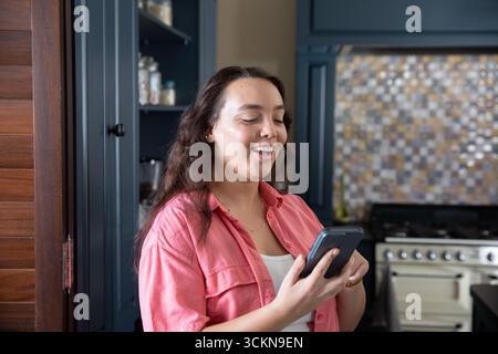 Frau mit Korallenhemd, Smartphone in der Hand, Lächeln in der Küche mit Gewürzgläsern und Backsplash Stockfoto