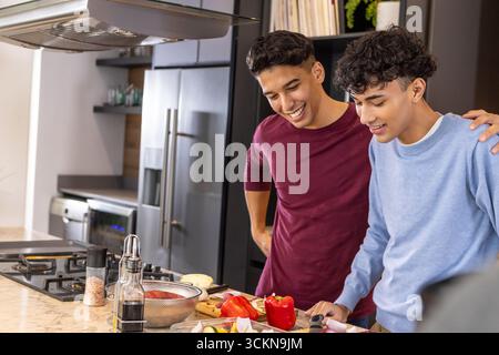 Zwei männliche Freunde hacken rote Paprika, Zucchini auf Marmorinsel mit Schneidebrett, Messer Stockfoto