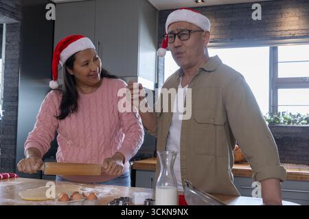 Verschiedene Paare in weihnachtsmannmützen bereiten an der Küchentheke Teig mit Nudelnadel und Eiern zu Stockfoto