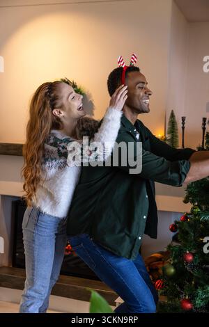 Verschiedene Paare, die weihnachtsbaum im Wohnzimmer mit Zuckerrohr, Stirnband und beleuchtetem Kamin dekorieren Stockfoto
