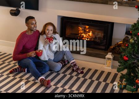 Diverse Paare sitzen auf gestreiften Teppichen vor dem Kamin am weihnachtsbaum mit roten Tassen Stockfoto
