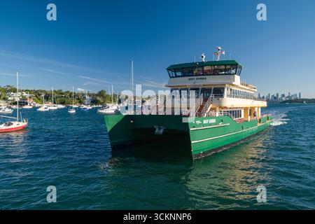 Eine Fähre namens May Gibbs fährt durch einen belebten Hafen voller vertäuter Boote, der vor einem lebhaften Hintergrund einer üppigen und üppigen Skyline der Küstenstadt liegt Stockfoto