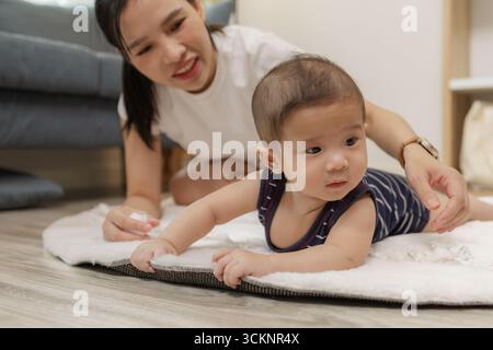 Ein Baby genießt die Bauchzeit auf einer weichen Matte, während die Mutter nah bleibt und es mit einem warmen Lächeln ermutigt. Dieser zärtliche Moment hebt frühe Entwicklungskräfte hervor Stockfoto