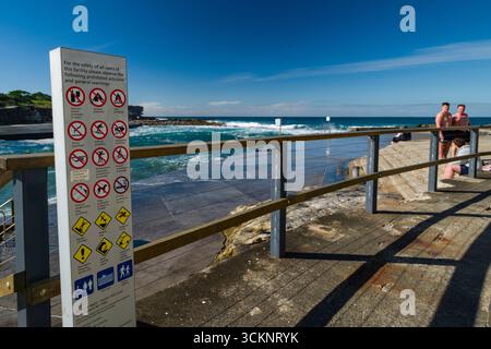 Ein landschaftlich schöner Pool im Pazifischen Ozean, eingerahmt von einem Geländer, mit einem auffälligen Warnschild. Besucher genießen die sonnige Atmosphäre am Wasser, während Waves cras Stockfoto