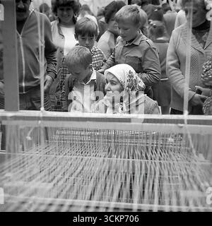 Eine Demonstration traditioneller Handwerkskunst während der Feierlichkeiten zum Stadttag in Sloviansk, 1988. Eine ältere Handwerkerin in einem traditionellen Kopftuch zeigt Kindern und Erwachsenen die Kunst des Webens auf einem alten Holzwebstuhl. Diese Szene spiegelt das starke Interesse an Volkskultur und nationalen Traditionen wider, das für die Perestroika-Ära charakteristisch war und die lebenswichtige Verbindung zwischen den Generationen und die Erhaltung des historischen Erbes in einer Zeit großer sozialer Veränderungen symbolisiert Stockfoto