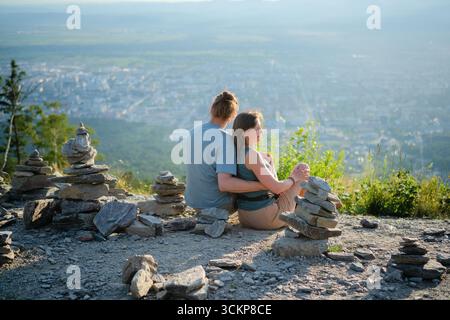 Mann und Frau sitzen von hinten umarmt und genießen während der Wanderung einen spektakulären Blick auf die Stadt, kunstvolle Steinstapel vorne. Reise- und Romantik-Thema. Stockfoto