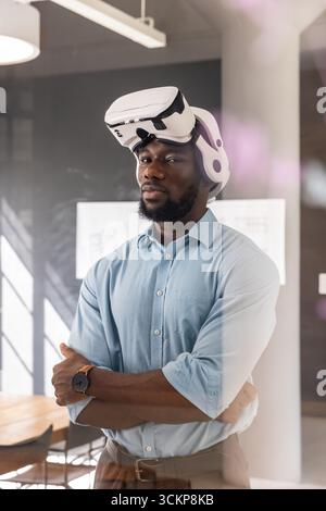 afroamerikanischer Mann mit vr-Headset, der mit überkreuzten Armen am Konferenztisch des Büros steht Stockfoto