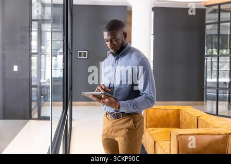 Afroamerikanischer Mann mit Business-Casual, der in der Lounge in der Nähe einer Glastrennwand steht und eine Tablette hält Stockfoto