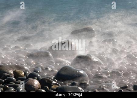 Wellen, die über die schwarzen Kieselsteine am Strand schweben Stockfoto